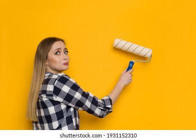 Sad And Scared Young Blond Woman In Lumberjack Shirt Is Painting Yellow Wall With Paint Roller And Looking At Camera Over The Shoulder. Waist Up Shot.