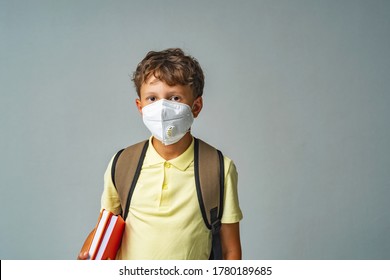 Sad Schoolboy With Backpack And Books In Protective Mask, Standing On Gray Background. Child Goes To School Upset Does Not Want To Wear A Mask. Protection During Epidemic Influenza And Coronaviruses