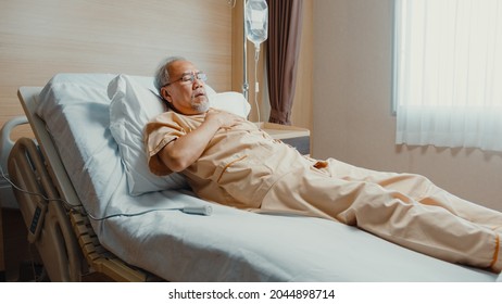 Sad senior Asia man having having heart attack lying on hospital bed and press emergency button. Sick aged guy lying hospitalized in a medical clinic. Health insurance or hospitality concept.