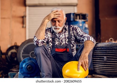 A sad senior factory worker sits next to the machines with a helmet in his hands and holding his head. He made a big mistake and he is afraid to be punished. The worker is worried.