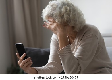 Sad senior lady using mobile phone at home, sitting on couch, covering face with hand. Frustrated upset mature 70s woman holding smartphone, reading message, making video call, getting bad news.