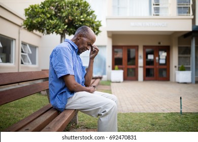 Sad Senior Man Sitting On Wooden Bench Outside Nursing Home