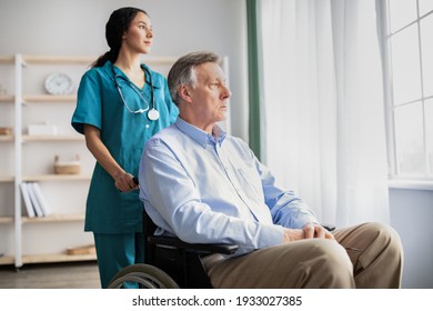 Sad senior man in wheelchair feeling depressed, looking out window, young nurse helping him at home. Caregiver assisting elderly male patient, taking care of pensioner at retirement facility