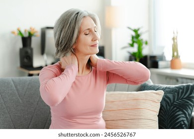 Sad senior woman having neck pain, sitting at sofa at home, with closed eyes. Health care concept - Powered by Shutterstock