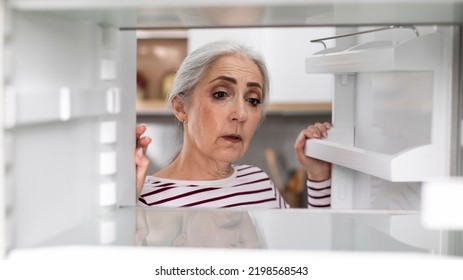 Sad Senior Woman Looking At Empty Shelves In Fridge At Home, Upset Elderly Lady Searching For Food In Refrigerator, Hungry Female Suffering Financial Crisis And Absence Of Money, View From Inside