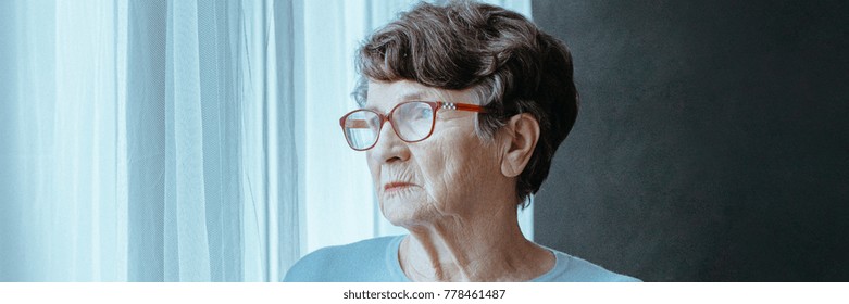 Sad Senior Woman Looking Through The Window Waiting For A Visit From Her Family, Feeling Lost And Lonely