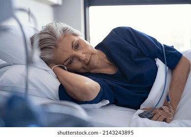 Sad senior woman lying on side on hospital bed. Depressed old patient lying on gurney at clinic with iv drip on hand. Elderly lonely woman with terminal illness resting at hospital ward. - Shutterstock ID 2143243895