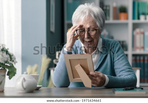Sad senior woman mourning the loss of\
her husband, she is holding a picture and\
crying