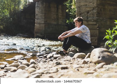 Sad Senior Woman Sitting Alone Near A River On A Summer Day - Depressed And Lonely Old Lady Looking In The Distance In Nature - Concept Image For Abandon And Loneliness