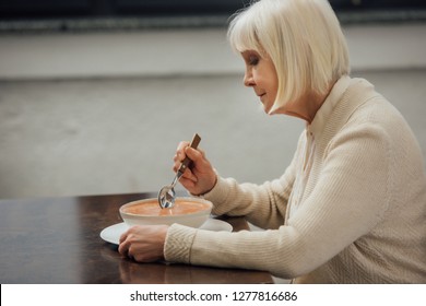 Sad Senior Woman Sitting At Table And Eating Cream Soup At Home