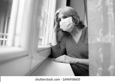 Sad Senior Woman Wearing Surgical Mask Looking Through The Window, Covid19 Pandemic 