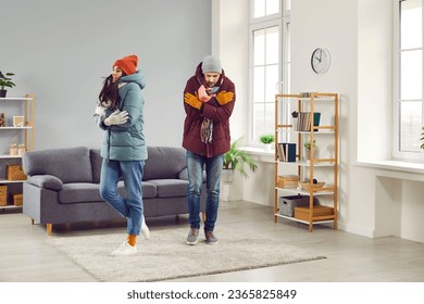 Sad shivering people in winter jackets freezing in cold apartment. Sad warmly dressed young man and woman feeling cold at home on winter season. Central heating problems concept