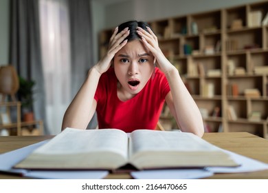 Sad Shocked Teen Chinese Lady Student With Open Mouth Study At Table With Book In Room Or Library Interior. Education At Home, Overwork, Exam Preparation And Facial Expression Due Covid-19 Pandemic