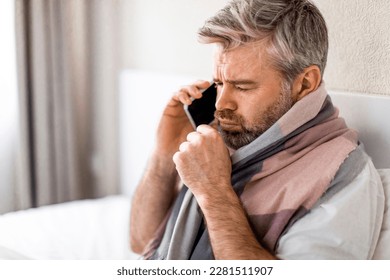 Sad sick middle aged grey-haired man with warm scarf around his neck sitting in bed at home, talking the phone to his employer while asking about the sick leave, calling doctor, coughing, copy space - Powered by Shutterstock