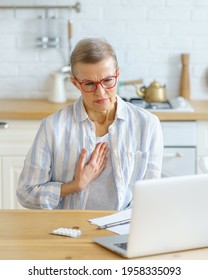 Sad Sick Middle Aged Woman Suffering From Pain In Chest Or Cough Having Online Consultation With Doctor On Laptop During Covid 19 Outbreak, Sitting In Kitchen At Home. Healthcare And Medicine Concept