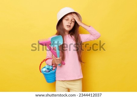Sad sick unhappy little cute girl in panama holding beach sandbox toys rake and shovel isolated over yellow background suffering heradache spending long hours under sun.