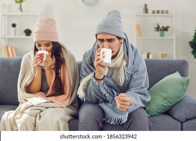 Sad sick young couple wrapped in plaid sitting on sofa in living-room drinking hot tea trying to warm up. Freezing man and woman suffering from cold or flu fever or having trouble with central heating