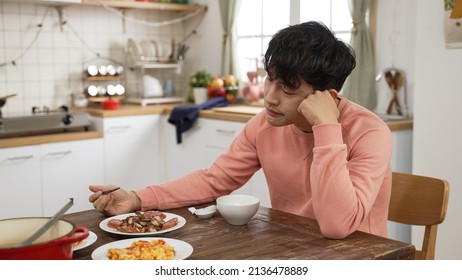 Sad Single Asian Male Having Lunch Alone During The Day In The Dining Room At Home. He Stops Eating And Props Head, Thinking About His Family And Friends.