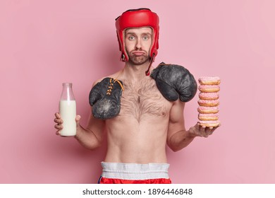 Sad Skinny Male Boxer Wears Hat And Boxing Groves Around Neck Holds Pile Of Donuts And Bottle Of Milk Poses With Naked Torso Isolated Over Pink Background Prepares For Battle. Sport Concept.