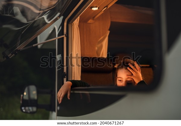Sad small girl by caravan window at dusk, family\
holiday trip.