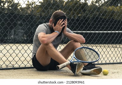 Sad sportsman tennis player sits on the court after lost match. Athlete man covers face with his hands.