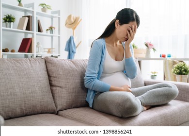 Sad And Stressed Pregnant Woman. Asian Young Female Lady At Home Depressed Not Ready To Be Mom Sitting On Couch Sofa At Home. Beautiful Upset Girl Cover Face By Hands Having Problem Parenthood