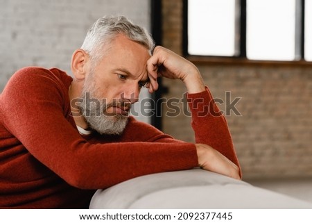 Sad stressed troubled mature middle-aged man sitting on the couch, missing his friends, feeling bad unwell unhealthy, suffering from depression at home