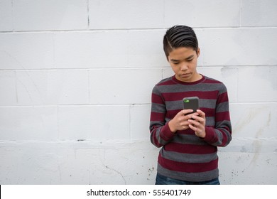 Sad teenage Asian boy standing alone against outdoor grungy white background using his smartphone.  Teen social concepts:  online bullying, peer pressure, social media competition, social networking