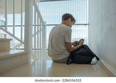 Sad teenage boy with mobile phone and backpack sitting on stairs. Teenager surfing on Internet, watching video, using app. Education, learning difficulties, mobile addiction concept - Powered by Shutterstock