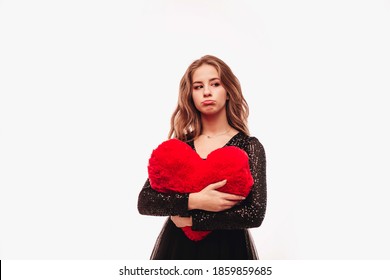 A Sad Teenage Girl With Curly Hair In Black Is Upset And Holding A Large Plush Heart, Isolated On White Background. Copy Space. 