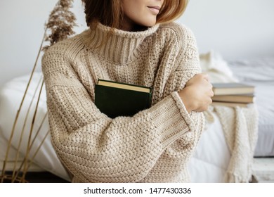 A sad teenage girl sits near bed in warm sweaters and holding book. Beige toned image - Shutterstock ID 1477430336