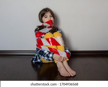 Sad Teenage Girl Sitting On The Floor, Tied With A Signal Tape. Quarantine, Self-isolation, Ban On Leaving The House. Child Protest, The Problem Of The Relationship Between Adolescents And Parents