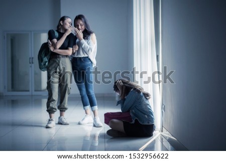 Sad teenage girl sitting on floor surrounded by classmates mocking her in the classroom