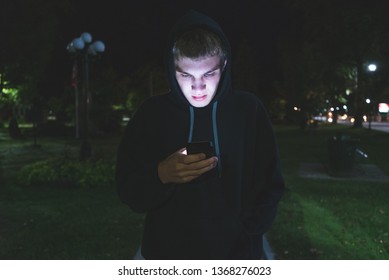 Sad Teenager On His Cellphone While Standing In A City Park At Night.