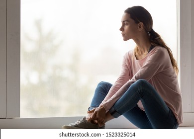 Sad thoughtful girl sit alone on sill looking through window feel pensive lonely, depressed melancholic upset young woman think of psychological problem regret mistake grieving in depression concept 