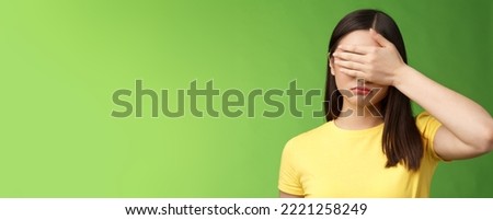 Sad tired asian girl dark haircut, shut eyes hold hand sight, grimacing, feeling gloomy and upset, stand green background depressed, unwilling watch how life ruins, pose yellow t-shirt.