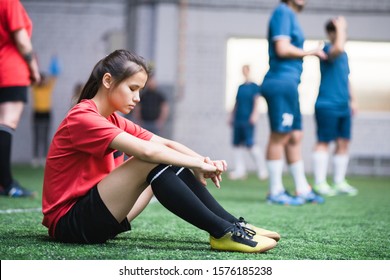 Sad or tired female football player in sports uniform sitting on green field on background of other team - Powered by Shutterstock