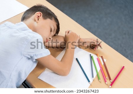 Sad tired frustrated boy lying on the table. Learning disability, reading difficulties, education concept. Top view of a primary school student who lies in front of a blank piece of paper with pencils
