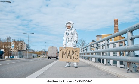 Sad And Tired Man In Spacesuit Is Standing At The Edge Of A Road And Holding A Sign With Mars Written On It. Astronaut Looking To Hitchhike A Car. Spaceman In Futuristic Suit