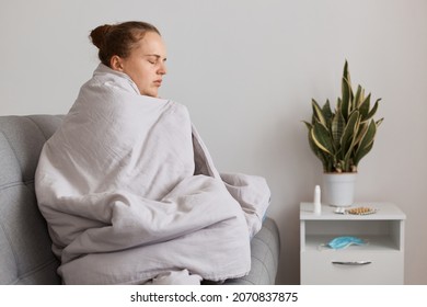 Sad tired woman with hair bun sitting on sofa wrapped in blanket felling very bed, having covid symptoms, having high temperature, fells cold, seasonal influenza.