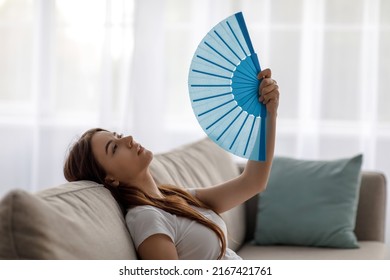 Sad Tired Young European Woman Suffering From Heat On Sofa And Waving Fan At Herself In Living Room Interior, At Window Background, Profile. Summer Weather And Overheating Without Conditioning At Home