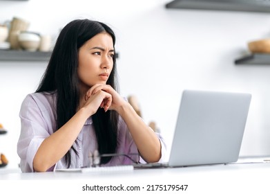 Sad unhappy Asian millennial girl, student or freelancer, sits at the table with a laptop, upset by a bad message, dissatisfied with the result of the project, did not receive approval, in a bad mood