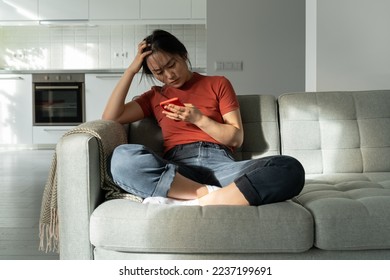 Sad unhappy Asian woman holding smartphone reading unpleasant message, breaking up over phone, sitting on sofa at home. Depressed upset girl receiving break up text from boyfriend. Unrequited love