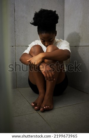 Sad, unhappy and depressed student sitting alone on the floor of a bathroom in school. Lonely, upset and depressed teen girl struggling with bullying and mental health in the corridor
