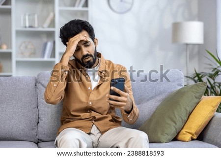 Sad unhappy and disappointed man sitting at home on sofa in living room, Indian man received notification message with bad news, reading using smartphone app.