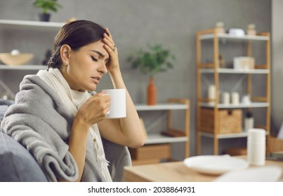 Sad unhappy exhausted sick woman suffering from common cold or flu fever, sitting on sofa wrapped in scarf, plaid and blanket, feeling unwell, touching forehead, holding mug and drinking warm tea