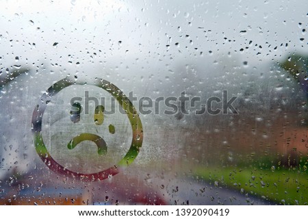 Sad unhappy face drawn on fogged glass on a wet rainy grey window soft light grey skies