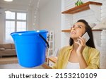 Sad unhappy frustrated young housewife lady looking up at water falling down from damaged ceiling in newly renovated home interior, holding pail and speaking to repair service. House roof leak concept