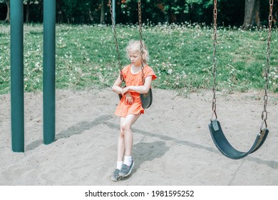 Sad Unhappy Girl Is Swinging On Swing Set In Park Outdoor Alone. Upset Bored Kid Waiting For Friend On Playground. Lonely Child Is Swinging Alone In Park And Missing Friend. Negative Emotion.