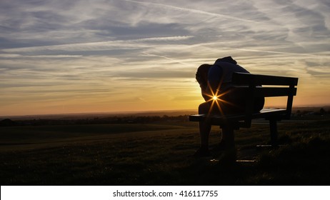 Sad unhappy silhouette man sitting with his head in his hands on a bench at sunset